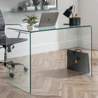 Amalfi clear glass curved desk styled in a modern home office with a laptop, lamp, and desk chair on a herringbone floor