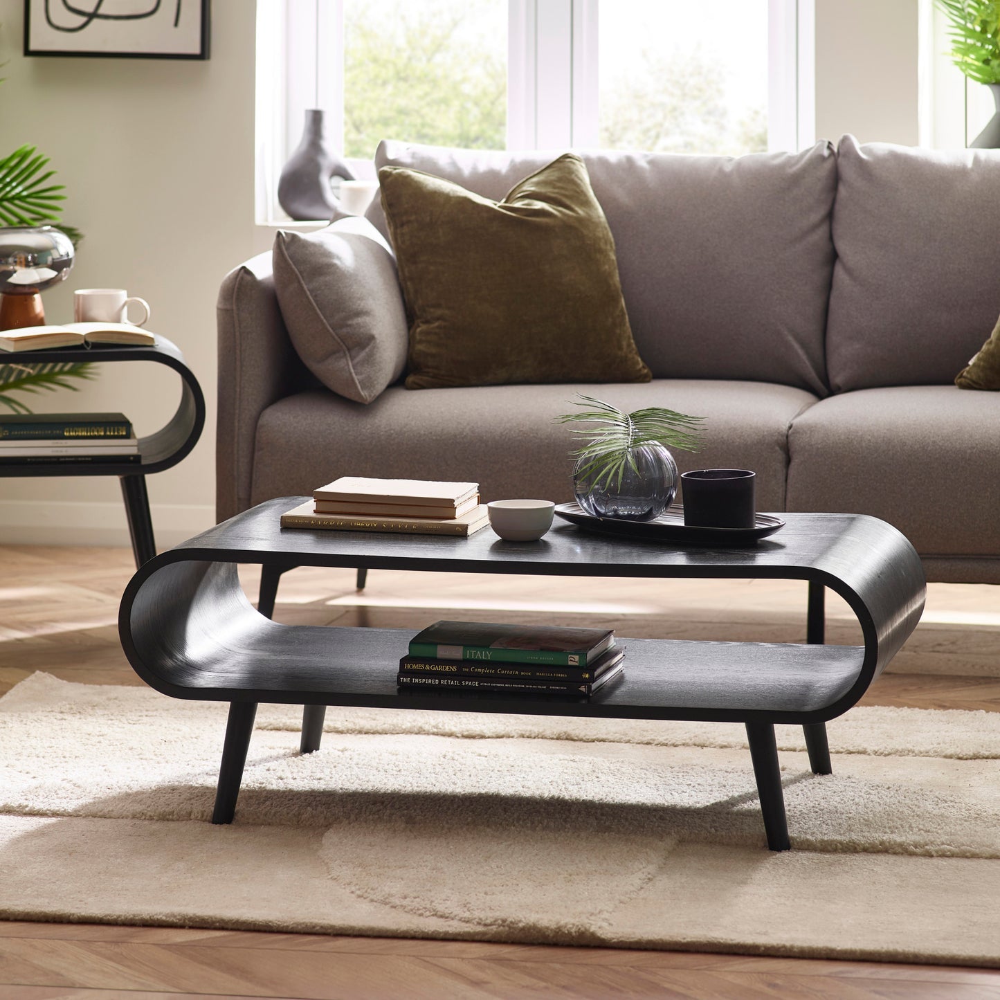Apollo black curved wooden coffee table styled with books and decor in front of a grey sofa on a neutral rug