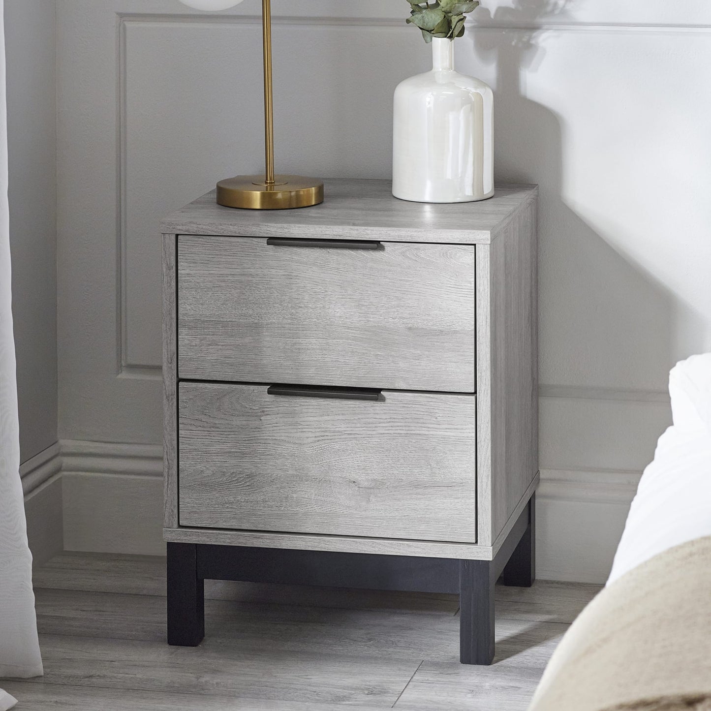 Bali grey oak effect 2 drawer bedside table styled with a lamp and vase in a bright bedroom setup