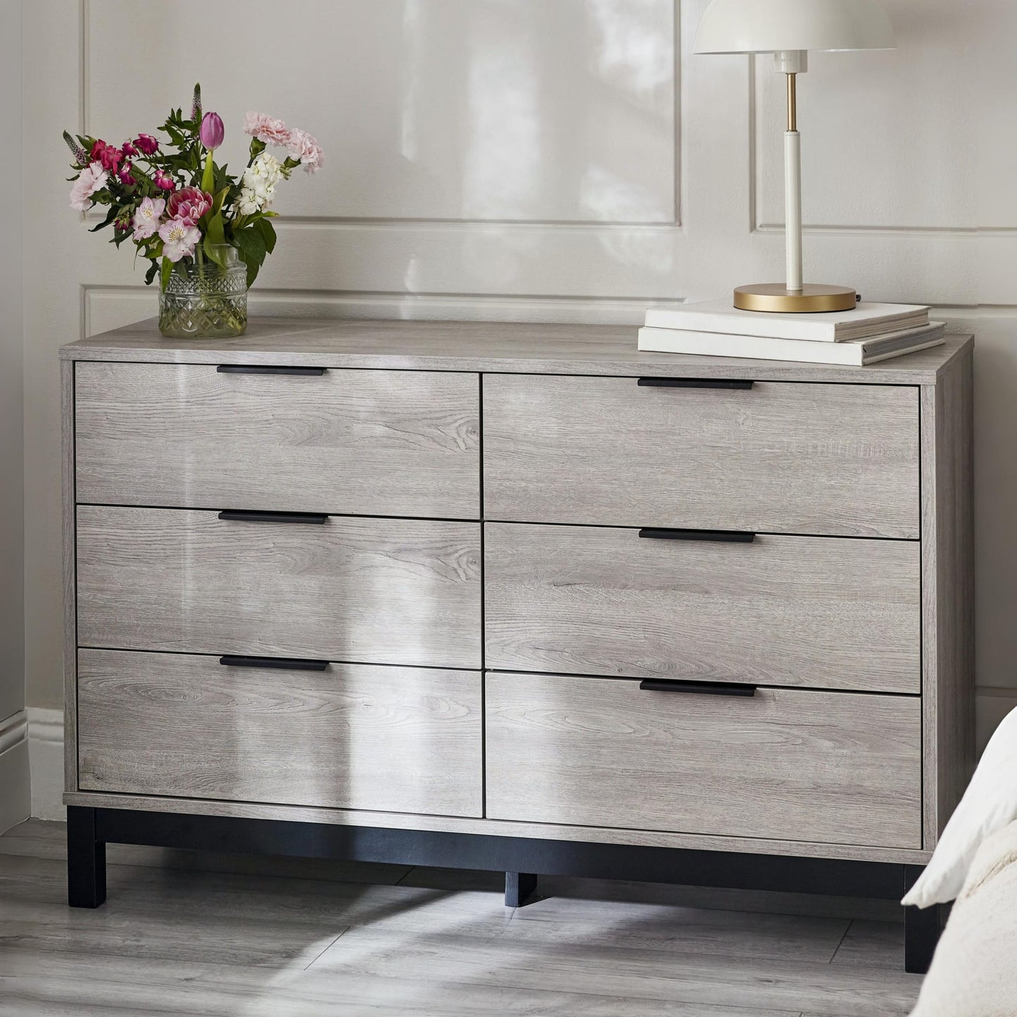 Bali grey oak effect 6 drawer wide chest styled in a bright, modern bedroom with flowers and a lamp