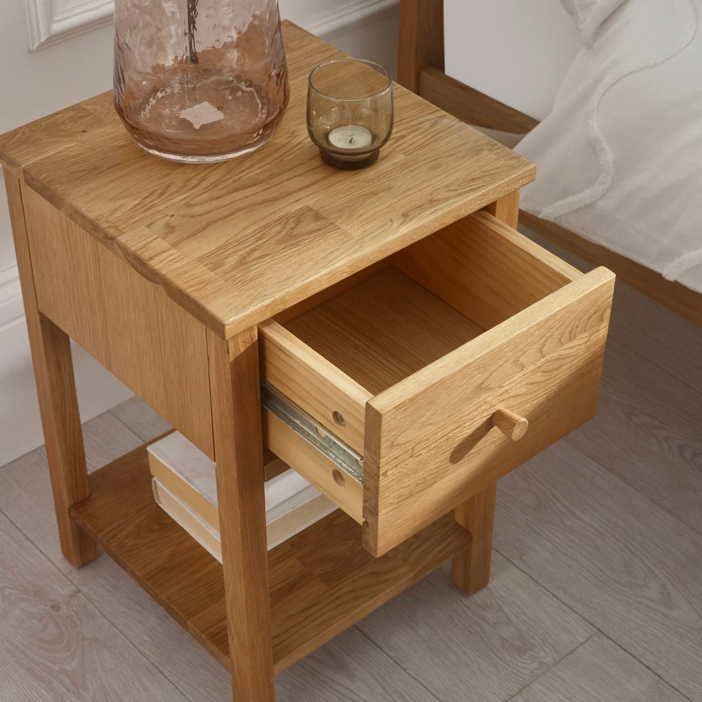 High-angle lifestyle shot of the Coxmoor oak nightstand with open drawer and lower shelf storage in a bedroom.