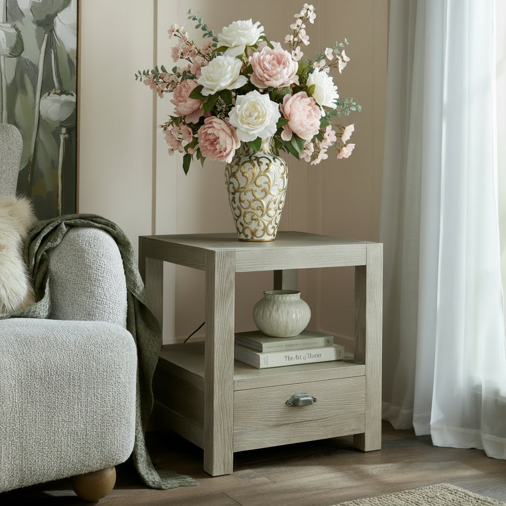 Haldon wooden 1 drawer side table with shelf with floral-patterned lamp and books next to a gray armchair in a room with white curtains.